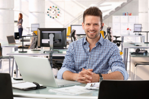 Happy casual successful caucasian business consultant at startup office, sitting at table with laptop computer, smiling, confident, looking at camera,